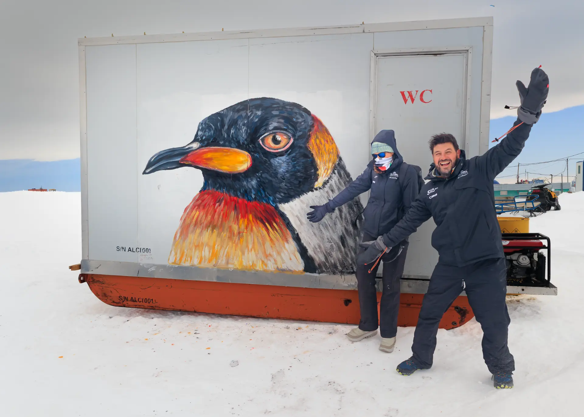 Two expedition members posing beside a large penguin mural painted on a container at camp