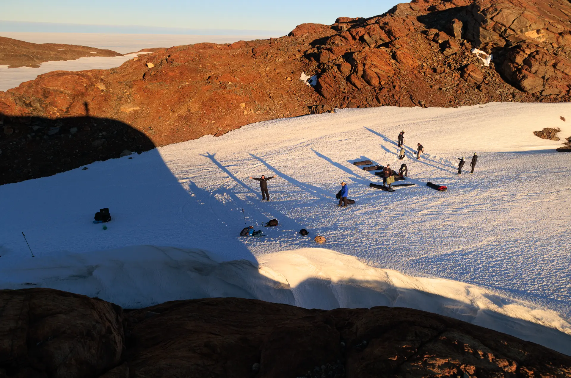 The expedition team spread across a sunlit snow slope below Antarctic rock