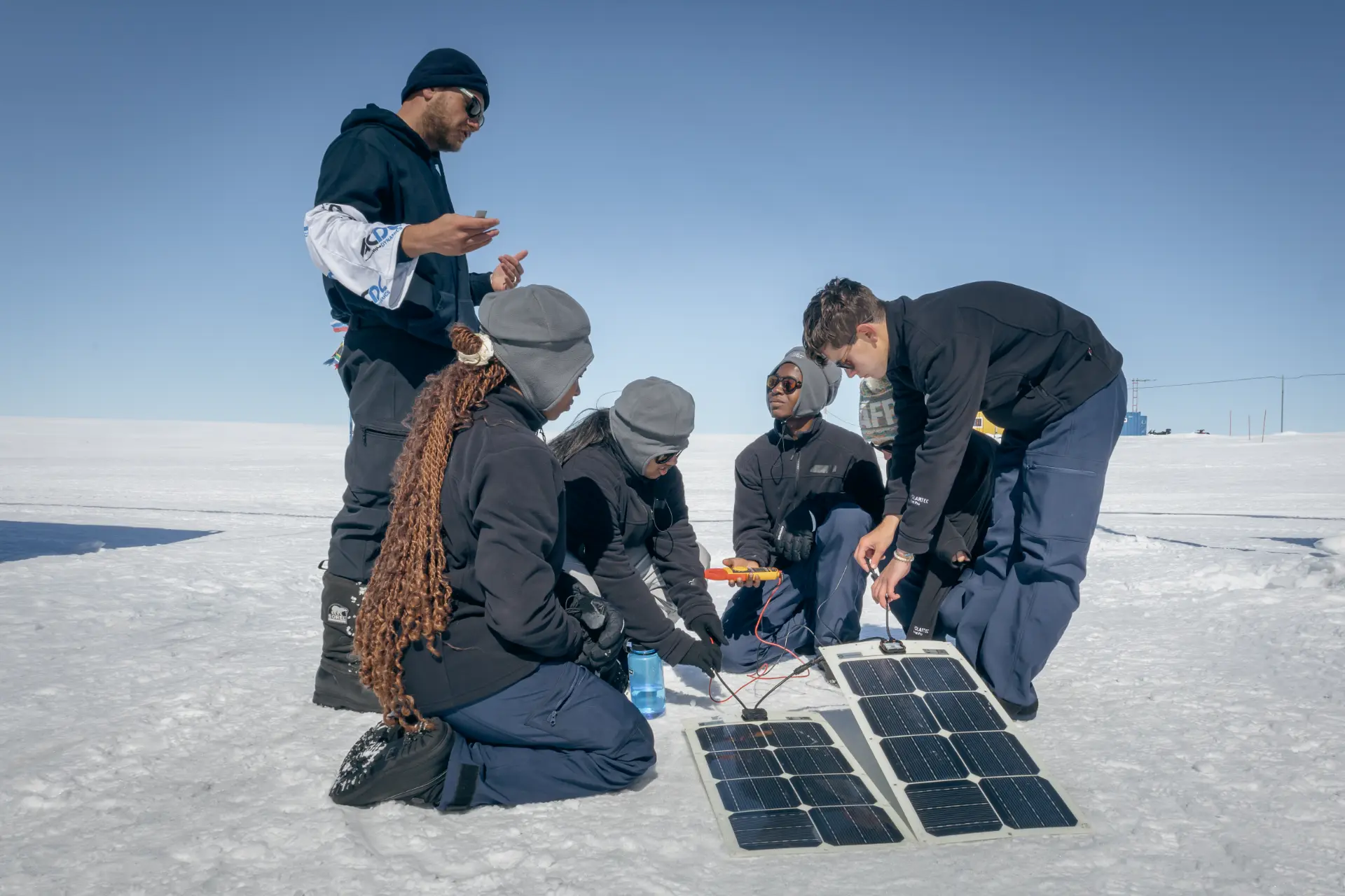 Six expedition members in cold weather gear kneeling on a bright Antarctic snowfield, testing portable solar panels and a multimeter together