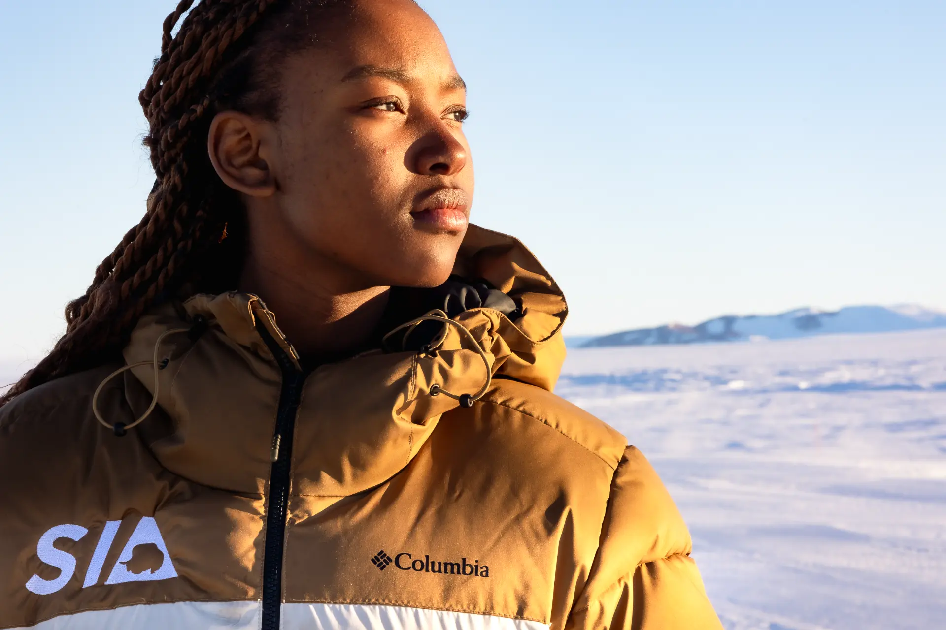 A student in a tan puffer jacket with an SIA patch looking thoughtfully toward the horizon against a bright out-of-focus snowy landscape