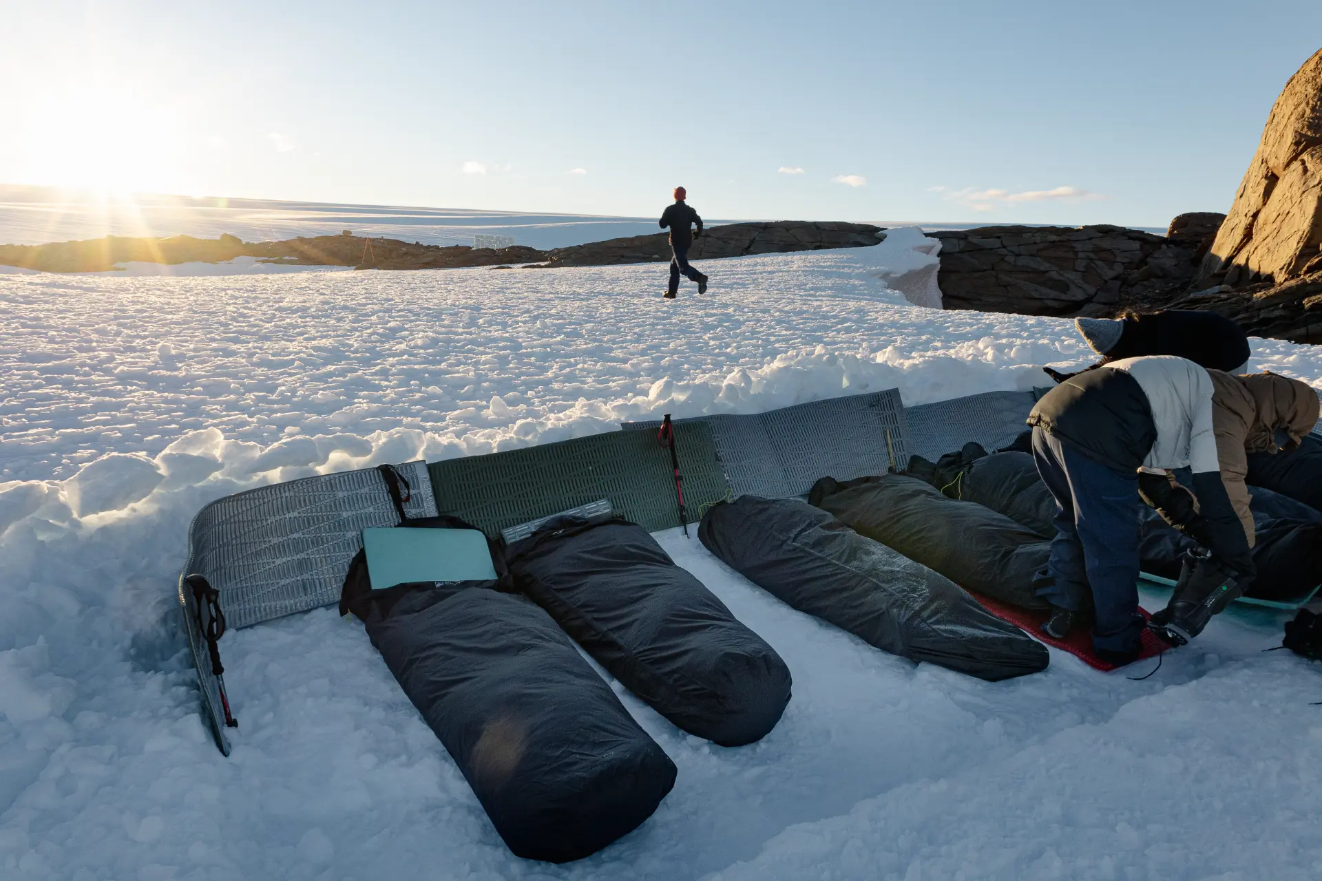 Sleeping bags and foam mats laid out across a vast sunlit Antarctic snowfield with expedition members tending to gear in the distance