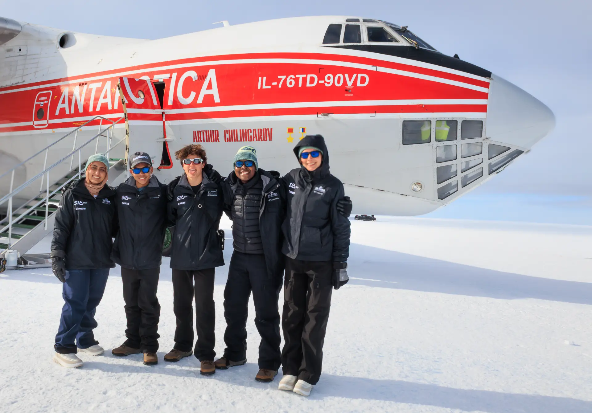 Five expedition members in black winter gear lined up in front of an Ilyushin cargo plane marked Antarctica on a bright Antarctic snowfield