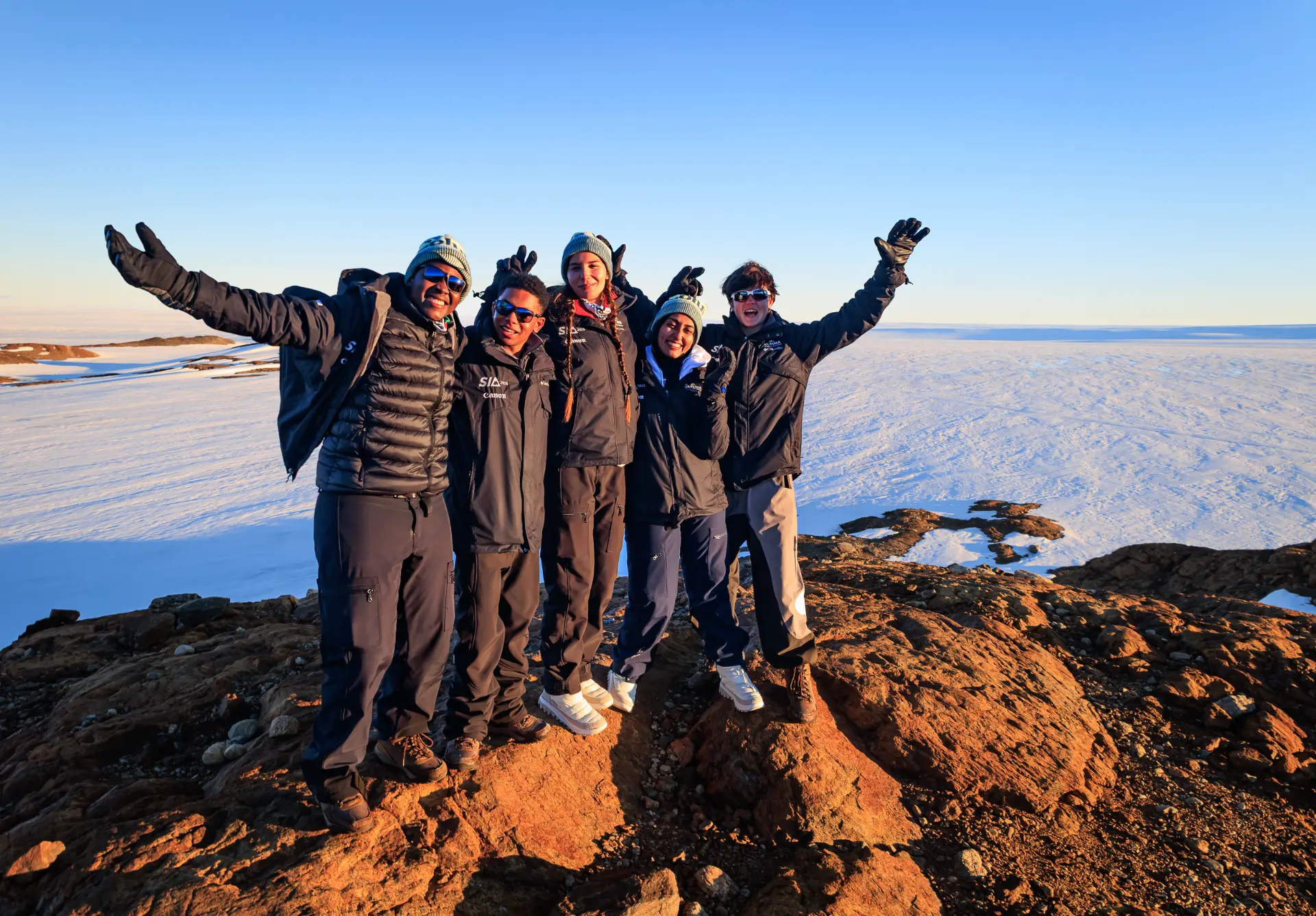 Five students standing on a sunlit rocky outcrop with arms raised above the Antarctic ice