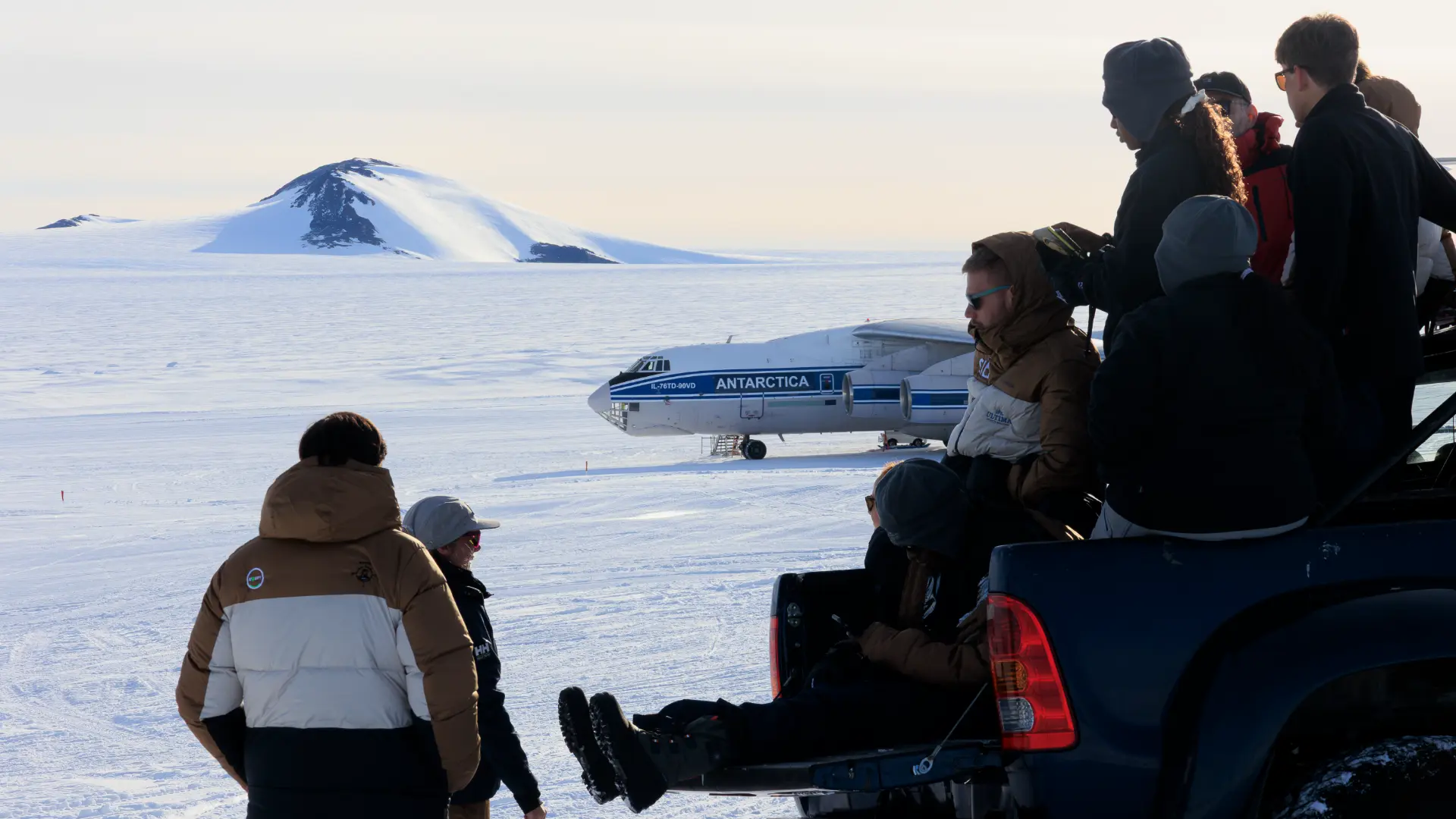 Expedition members on and around a vehicle watching the Ilyushin aircraft on the Antarctic runway