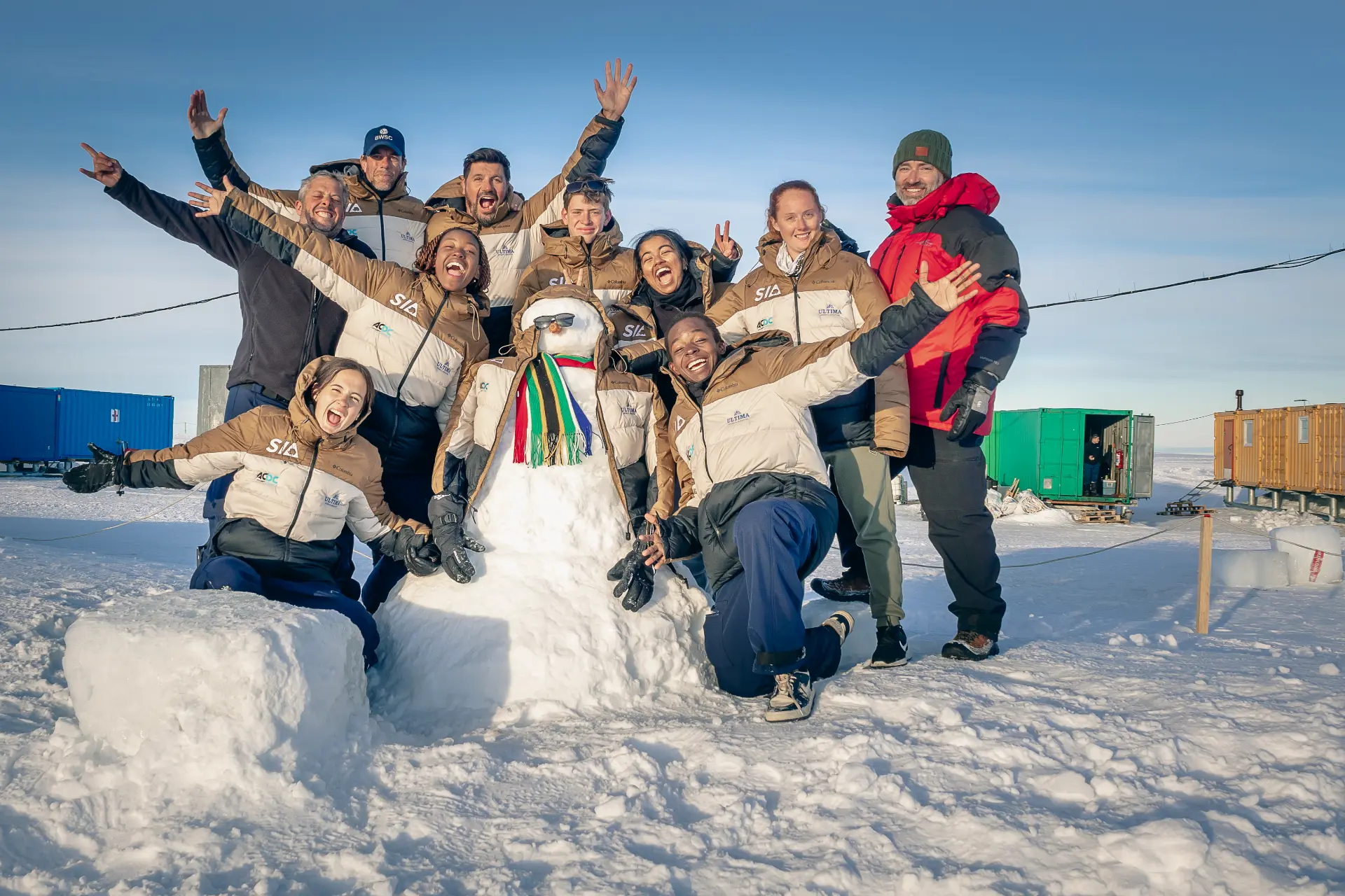 The expedition team gathered around a snowman wearing a scarf and sunglasses on the ice