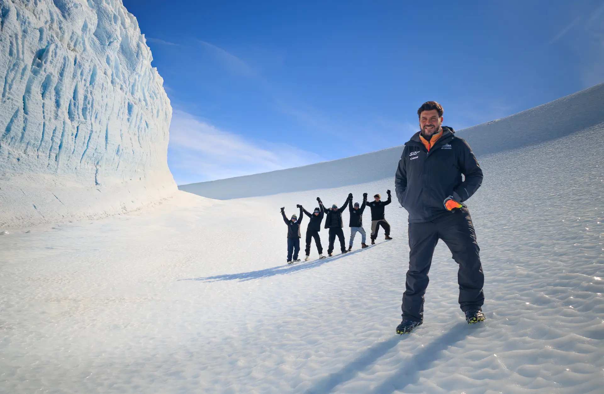 Riaan Manser in the foreground with five Grade 11 students behind him, arms raised triumphantly against an Antarctic ice wall. From left to right: Aaminah, Allegra, Ntokozo, Luke, Naethan.