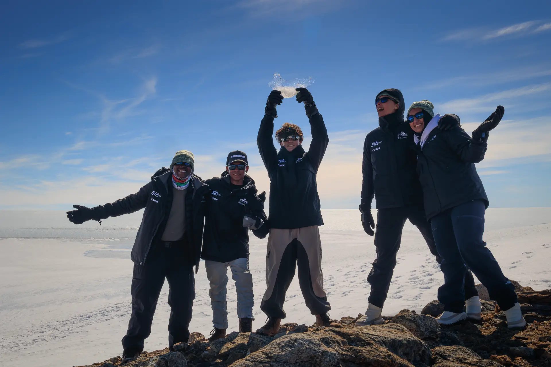 Students in Antarctica team celebrating with a shard of translucent ice raised overhead on the ice shelf